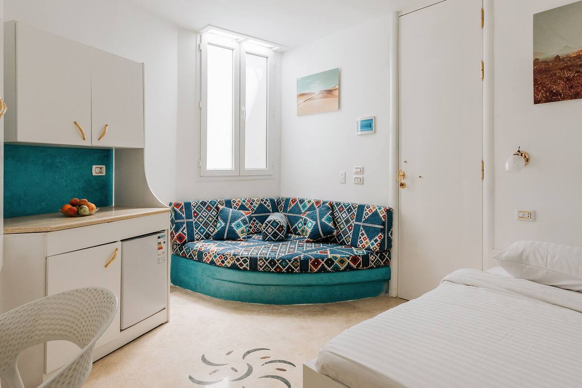Ein gemütliches Schlafzimmer mit einem Bett, einem Schreibtisch mit Stuhl, einem farbenfrohen Sofa und einem Fenster.