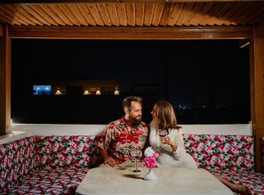 A couple seated on a floral-patterned sofa under a wooden ceiling, with a view of a building in the background.