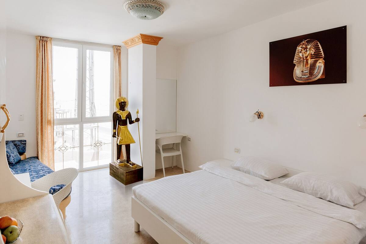 Ein Schlafzimmer mit einem Bett, einem Schreibtisch, einem Stuhl, einer Statue und einem Fenster mit Vorhängen.