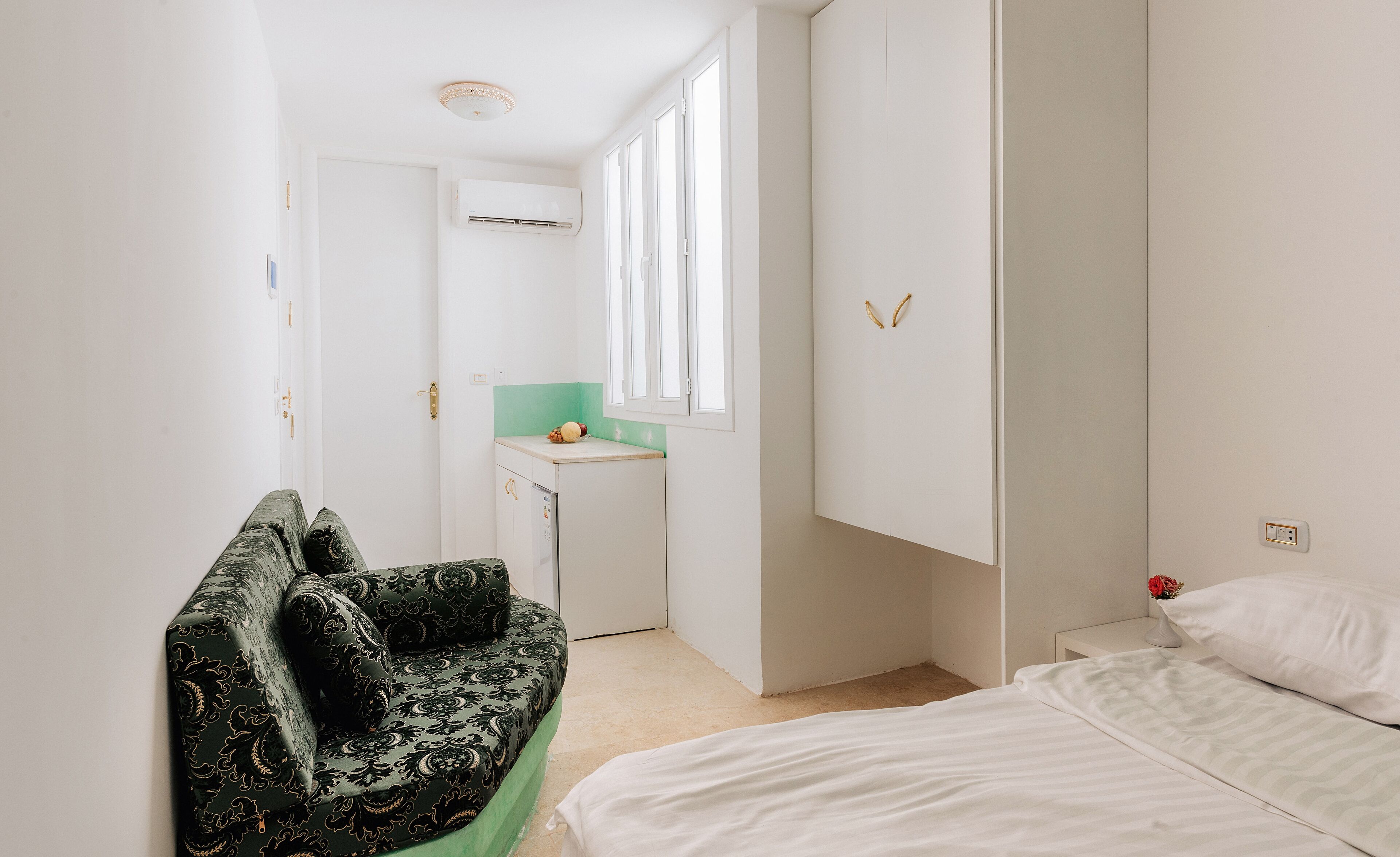 A compact bedroom with a bed, a green sofa, a small kitchenette, and a window.