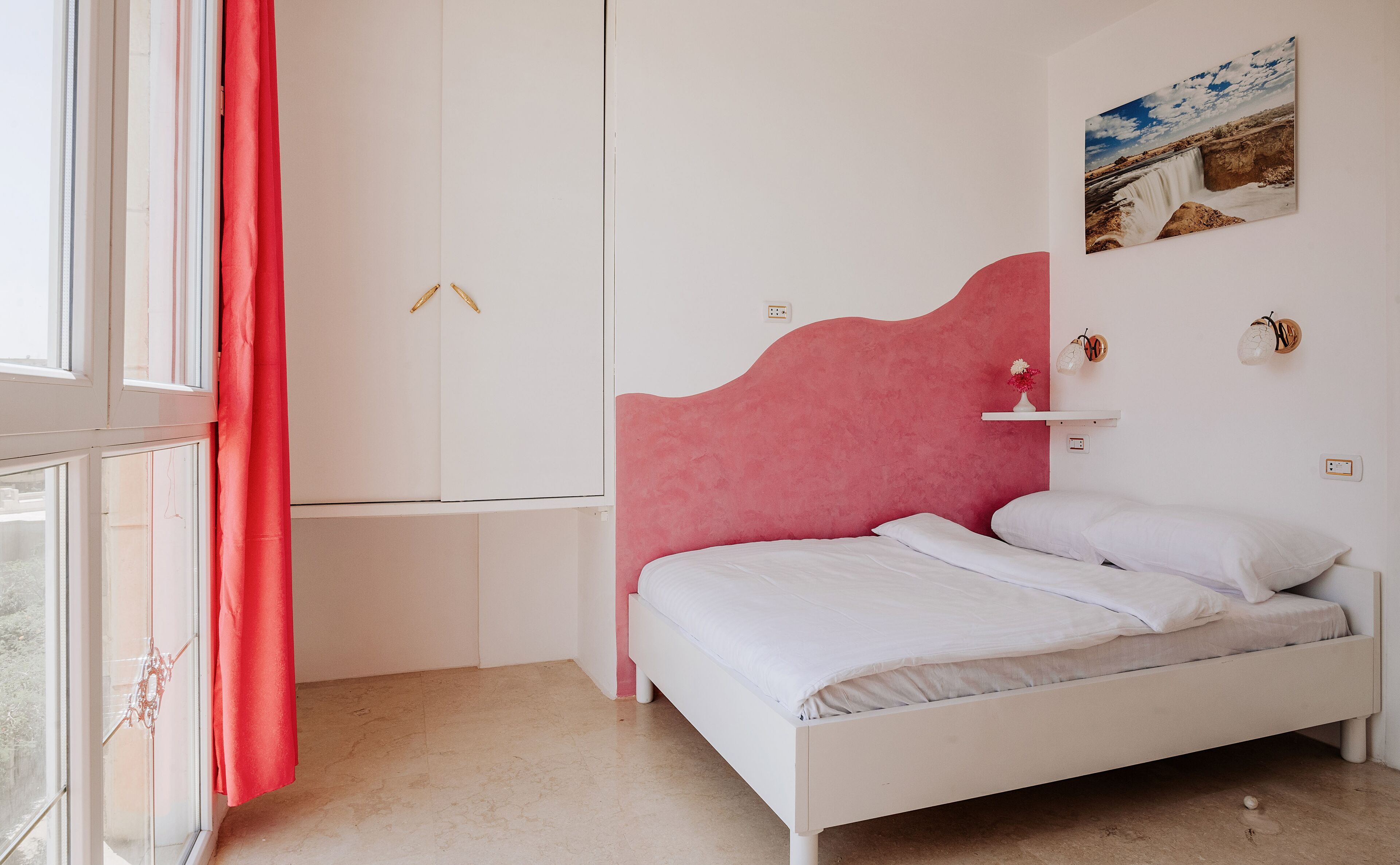 A bedroom with a bed, a wardrobe, a window, and a wall-mounted shelf with a vase.
