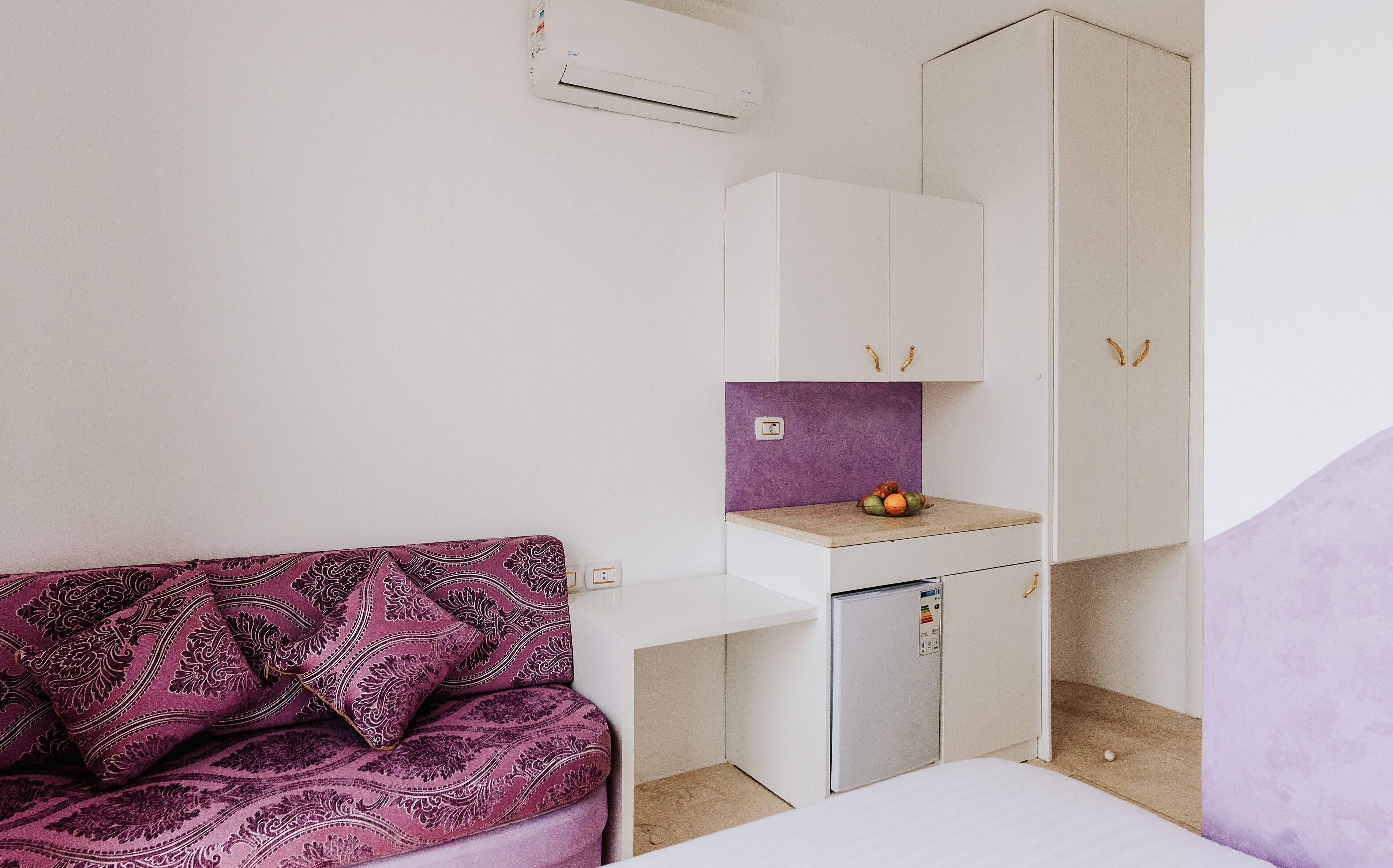 A small kitchenette with a refrigerator, a sink, and a purple backsplash. There is a white cabinet above the sink and a white wardrobe to the right. A purple patterned sofa is in the foreground.
