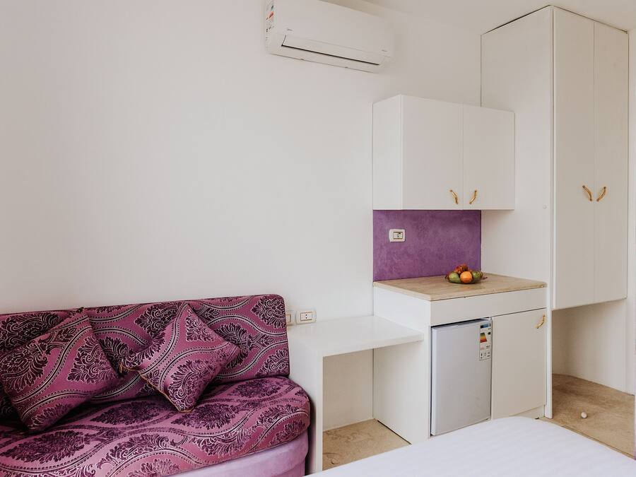 A small kitchenette with a refrigerator, a sink, and a purple backsplash. There is a white cabinet above the sink and a white wardrobe to the right. A purple patterned sofa is in the foreground.