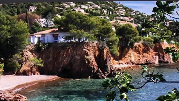 A coastal scene with clear turquoise water, rocky shore, and a hillside with buildings. - Apartment t2 type maisonette with garden sea view  (Theoule Sur Mer)