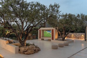 An outdoor seating area with a flat-screen TV, stone walls, and olive trees.