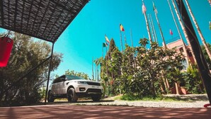 Ein weißer SUV parkt im Schatten, im Hintergrund Palmen und Flaggen.