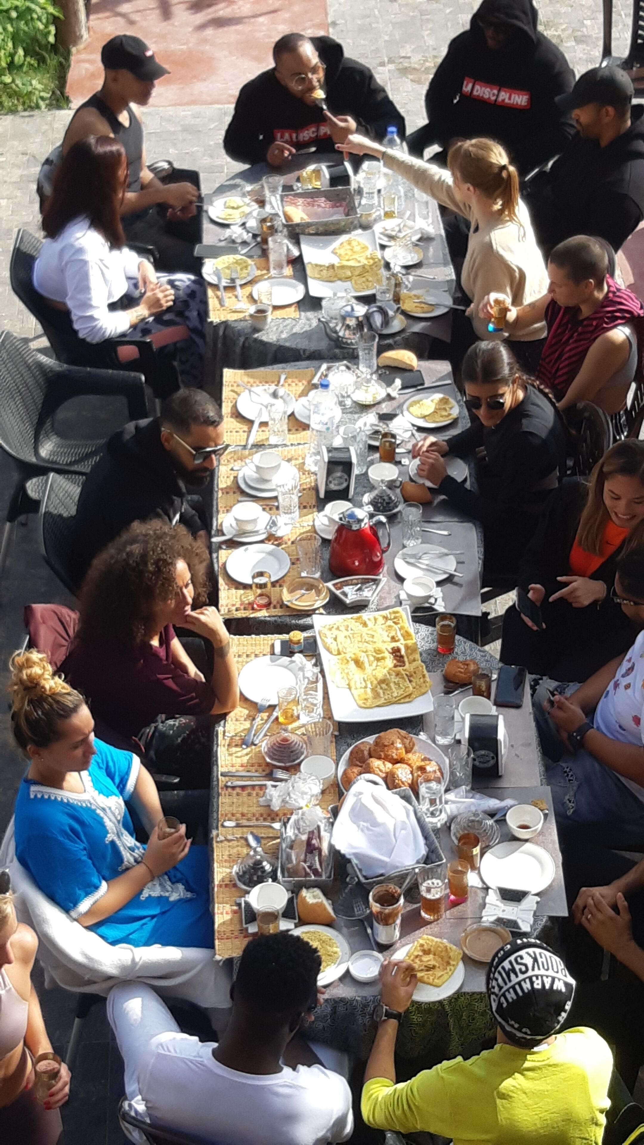 A group of people seated around a long table with various dishes and drinks, indicating a communal dining setting.
