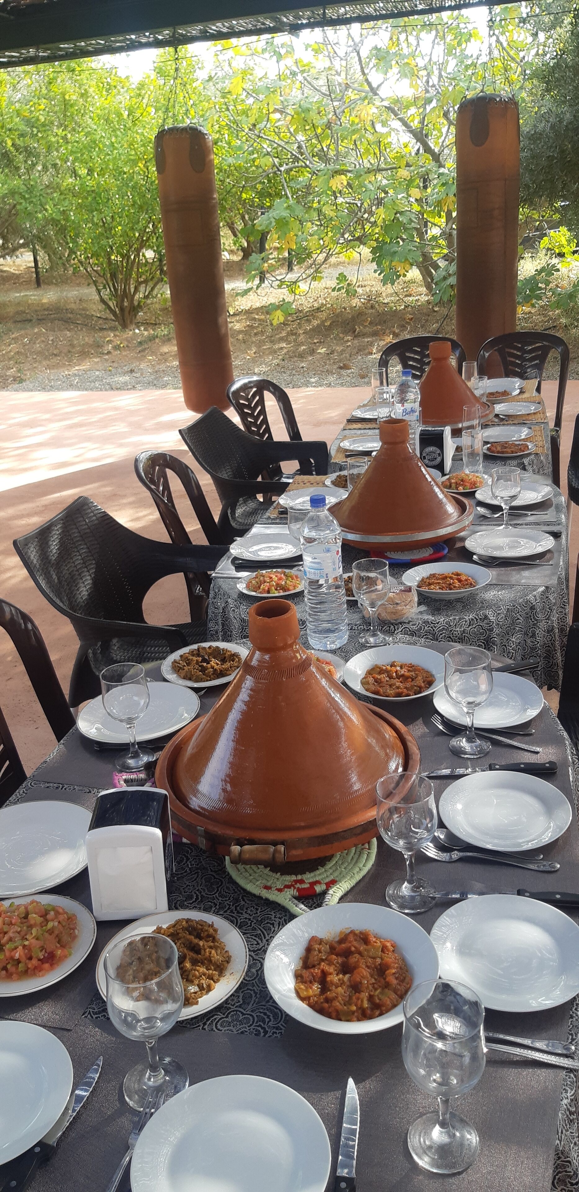Uma área de refeições ao ar livre com uma mesa posta para uma refeição, incluindo pratos, copos e uma grande panela de barro tradicional.