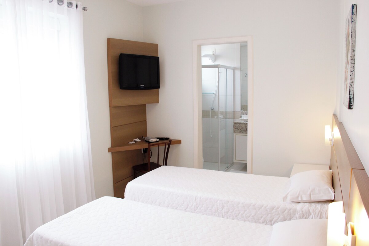 A hotel room with two beds, a TV mounted on a wooden panel, a desk with a chair, and a bathroom visible through a glass door.