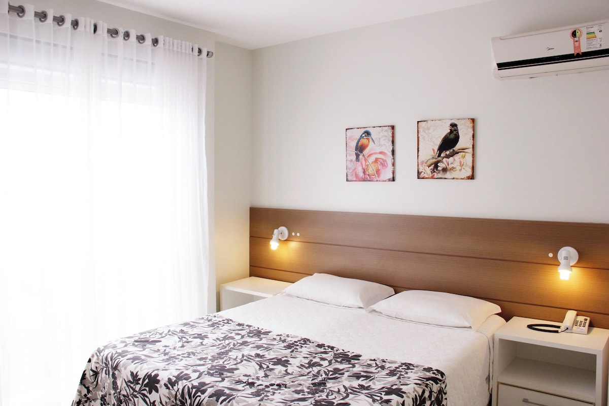 Habitación de hotel con cama, cortinas blancas, dos lámparas fijadas a la pared, aire acondicionado mural y dos cuadros enmarcados.