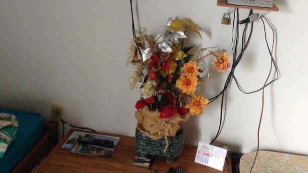 Una habitación con un banco de madera, un televisor de pantalla plana montado en la pared, un estante pequeño con un módem y un florero con flores sobre el banco.