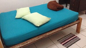 Una cama individual con colchón azul y dos almohadas, un cojín marrón, cabecero de madera, mesita de noche de madera oscura con una lámpara y una alfombra estampada sobre suelo de baldosas.
