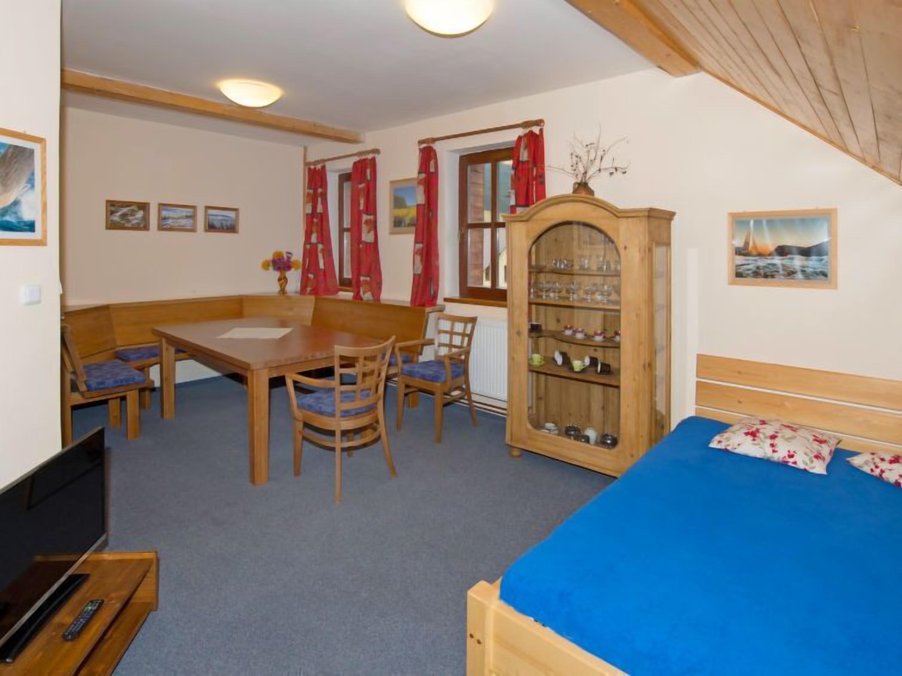 A cozy room with a wooden dining table, chairs, a bed with a blue mattress, and a cabinet with glass doors.