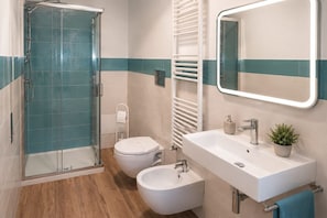 A bathroom with a glass shower enclosure, a toilet, a bidet, a sink with a mirror, and a towel rack.