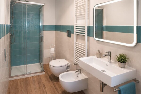 A bathroom with a glass shower enclosure, a toilet, a bidet, a sink with a mirror, and a towel rack.