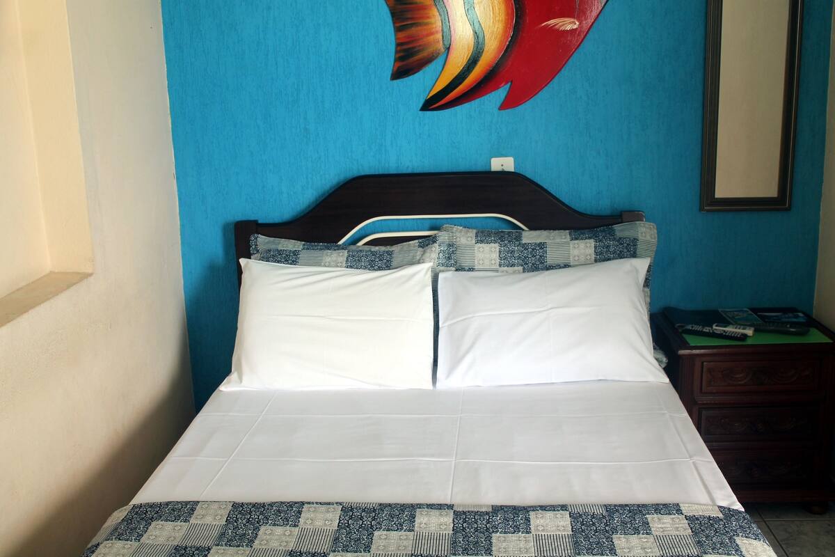 Ein Bett mit weißen Leinentüchern und einer gemusterten Tagesdecke, ein Holz-Kopfteil und ein Nachttisch mit einem Buch und einem grünen Gegenstand.