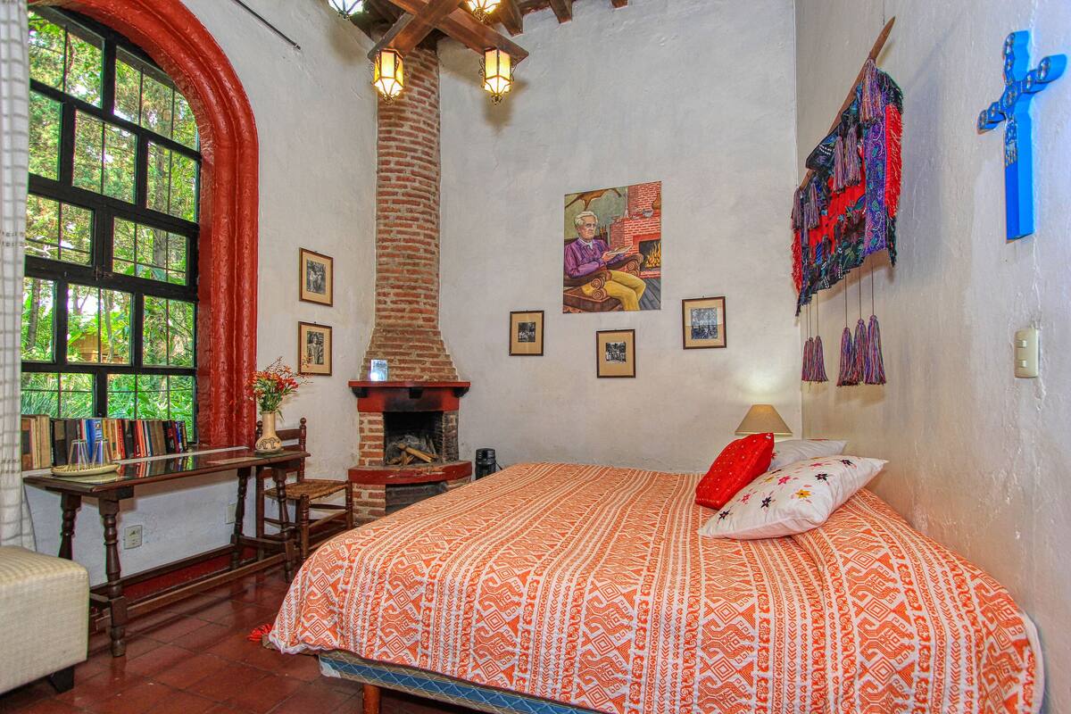 A room with a bed, a fireplace, a desk, and a large window.