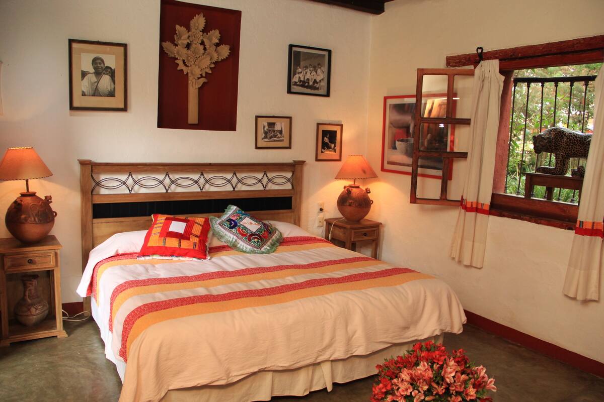 A neatly arranged bedroom with a bed, bedside lamps, a vase, and framed pictures on the wall.