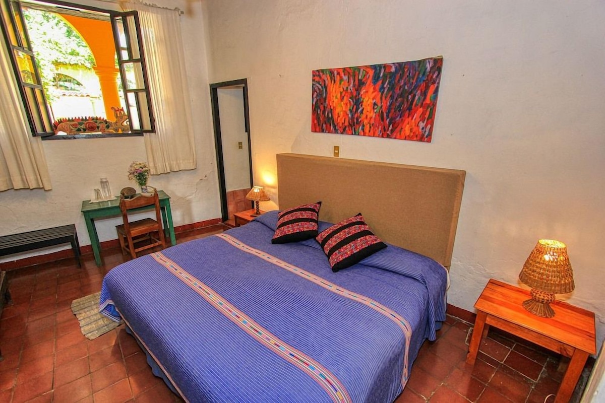 A bedroom with a bed, a nightstand, a lamp, a chair, a table, a mirror, and a window with curtains.