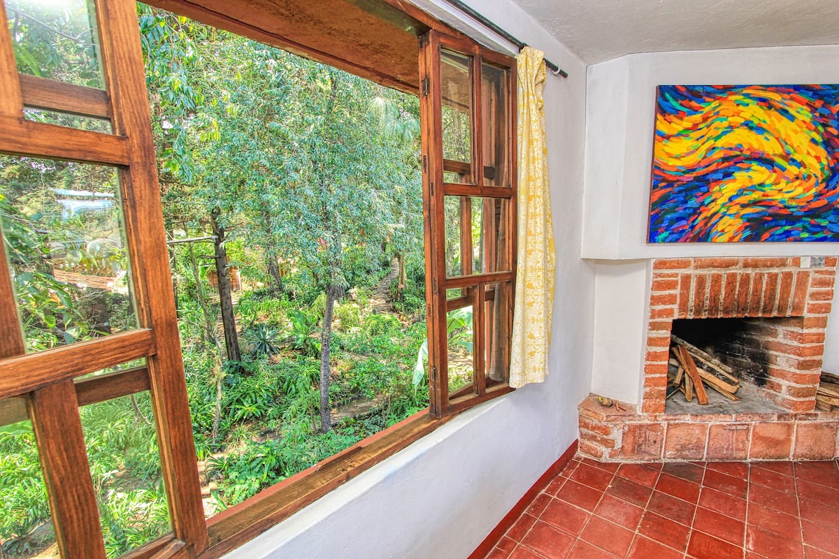 A room with a large window overlooking a lush garden, a brick fireplace, and a colorful abstract painting on the wall.