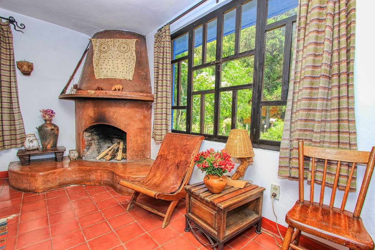 A rustic room with a wooden fireplace, a large window with curtains, and a wooden chair.