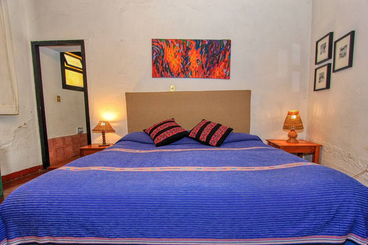 A bedroom with a large bed, a headboard, two bedside lamps, and a colorful abstract painting on the wall.