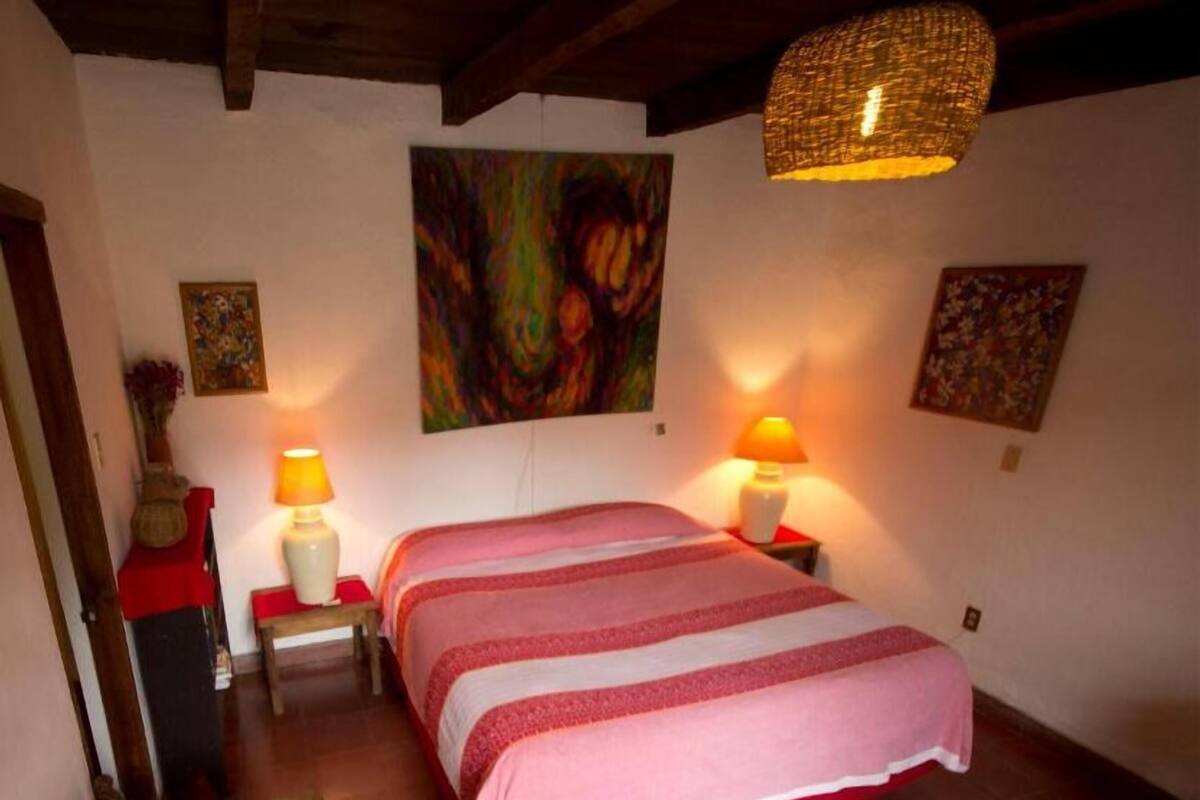 A bedroom with a bed, two bedside lamps, a painting on the wall, and a hanging light fixture.