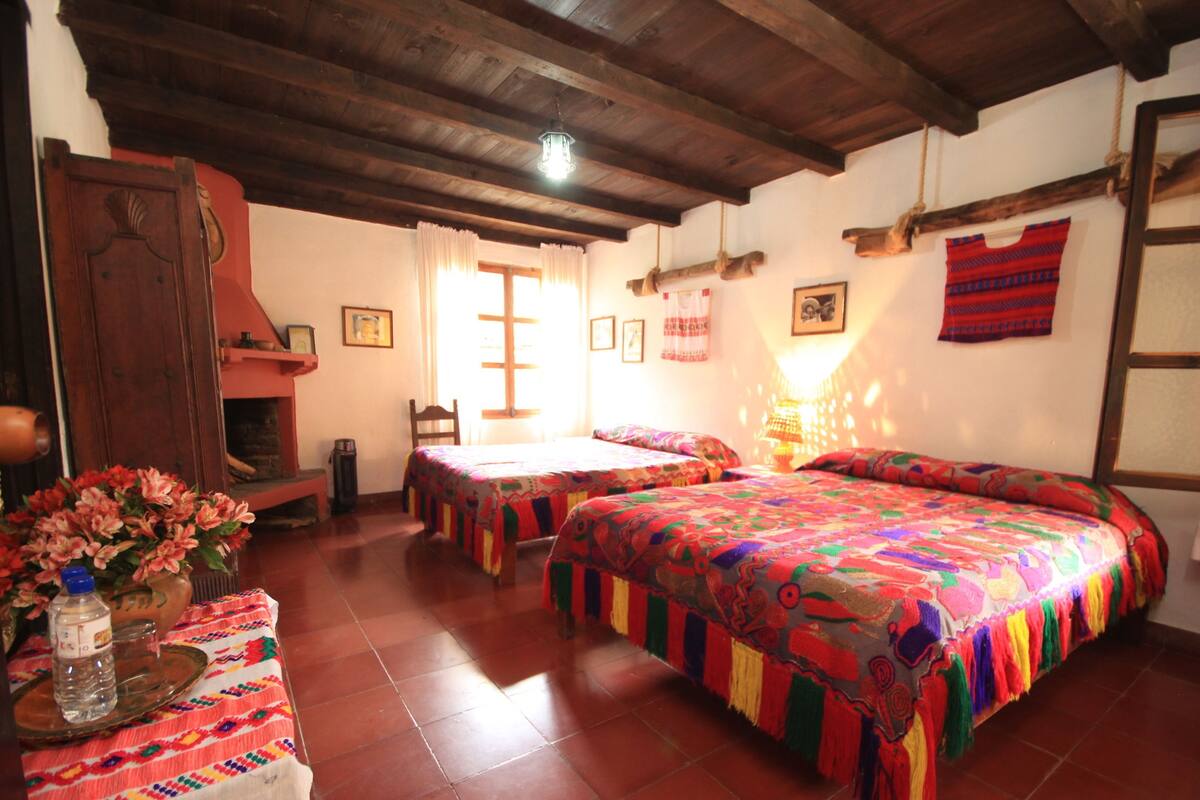 A room with wooden beams, a bed with colorful bedding, a wooden table with a vase, and a chair.