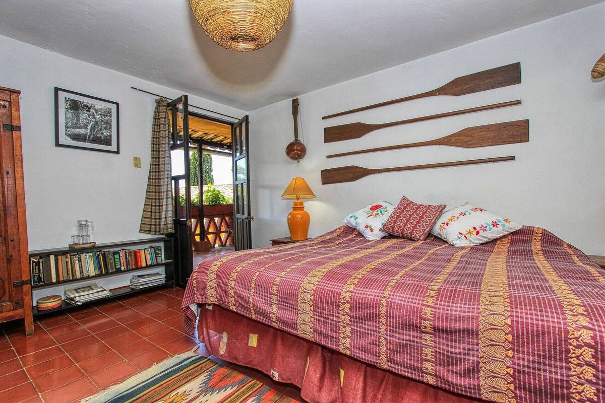 A room with a bed, a bookshelf, a wicker ceiling light, and a view through an open door.