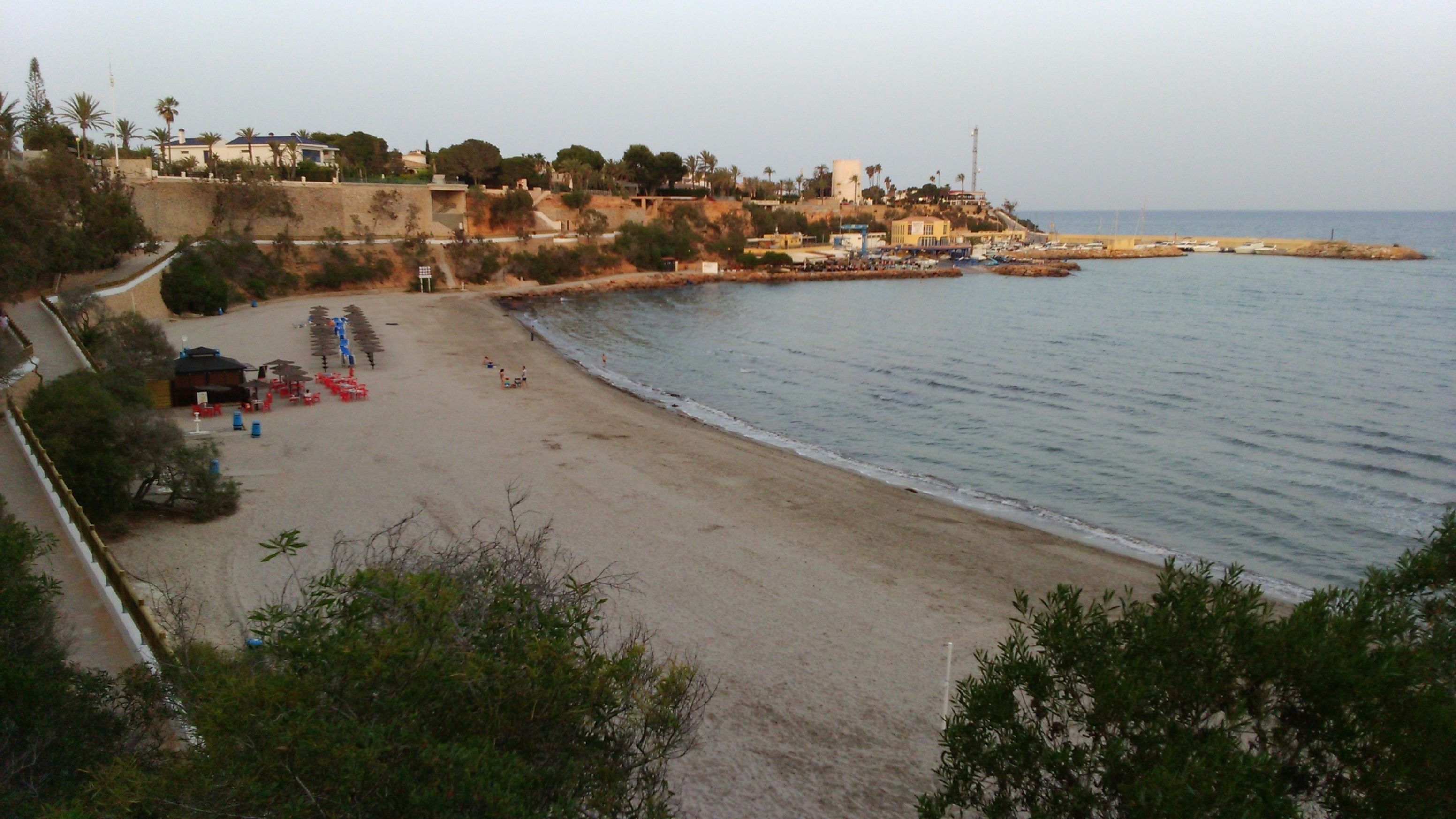 Une plage avec un rivage sablonneux, quelques parasols et quelques personnes. On aperçoit des bâtiments au loin et un phare sur la droite.