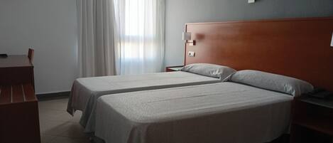Habitación de hotel con dos camas, cabecera de madera, mesita de noche y escritorio con lámpara.
