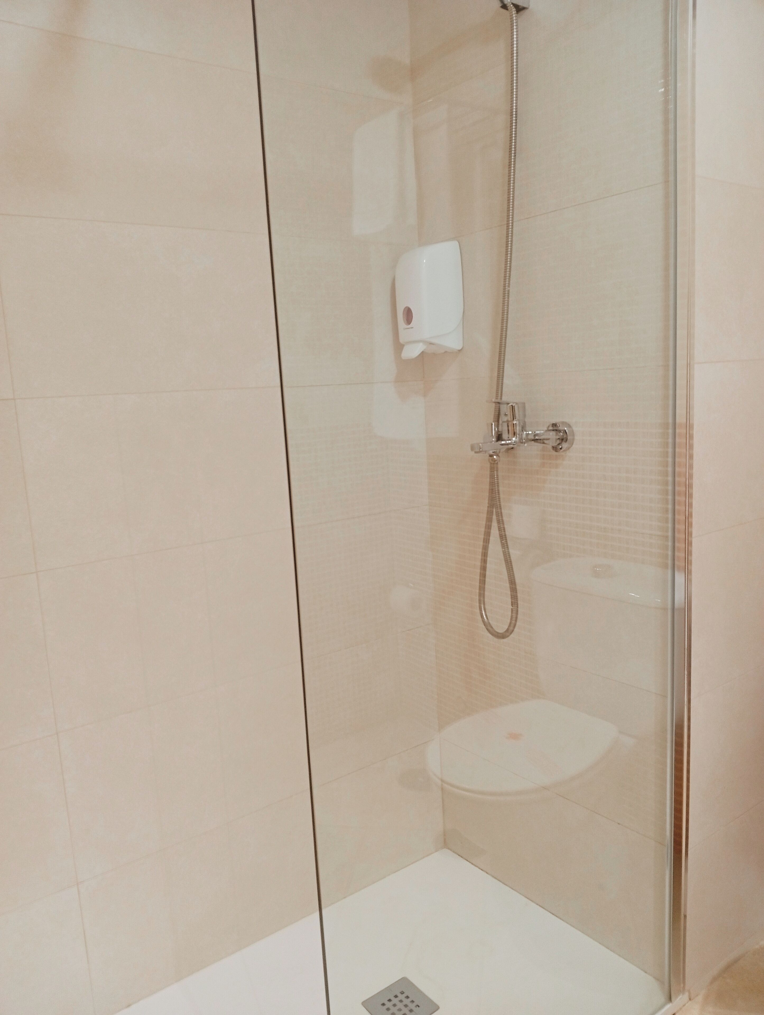 A glass-enclosed shower stall with a handheld showerhead and a soap dispenser on the wall.
