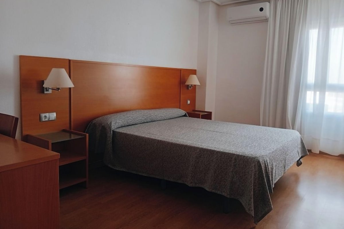 A hotel room with a bed, two bedside tables, a desk, and a window with curtains.