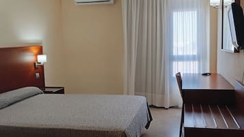 A hotel room with a bed, a desk, a chair, a TV, and an air conditioning unit.