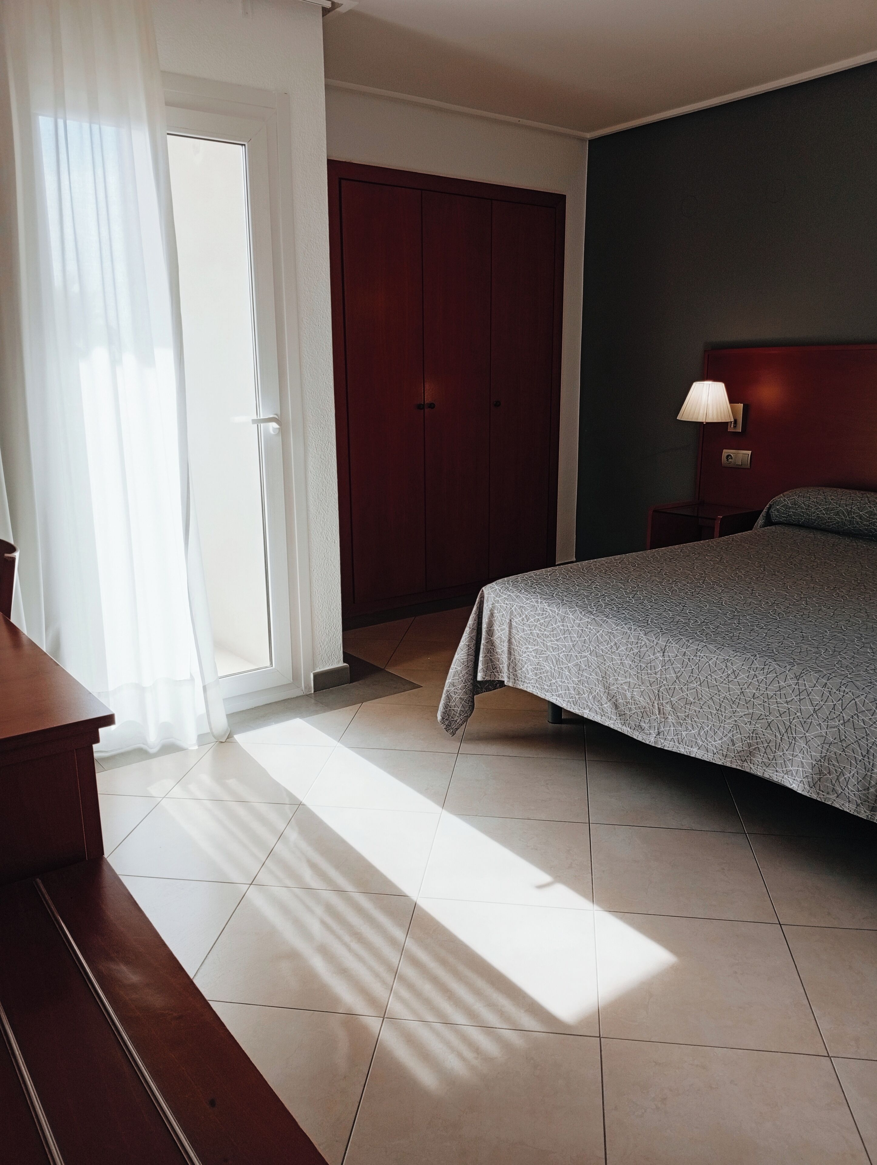 A hotel room with a bed, a nightstand, a wooden wardrobe, and a window with curtains.