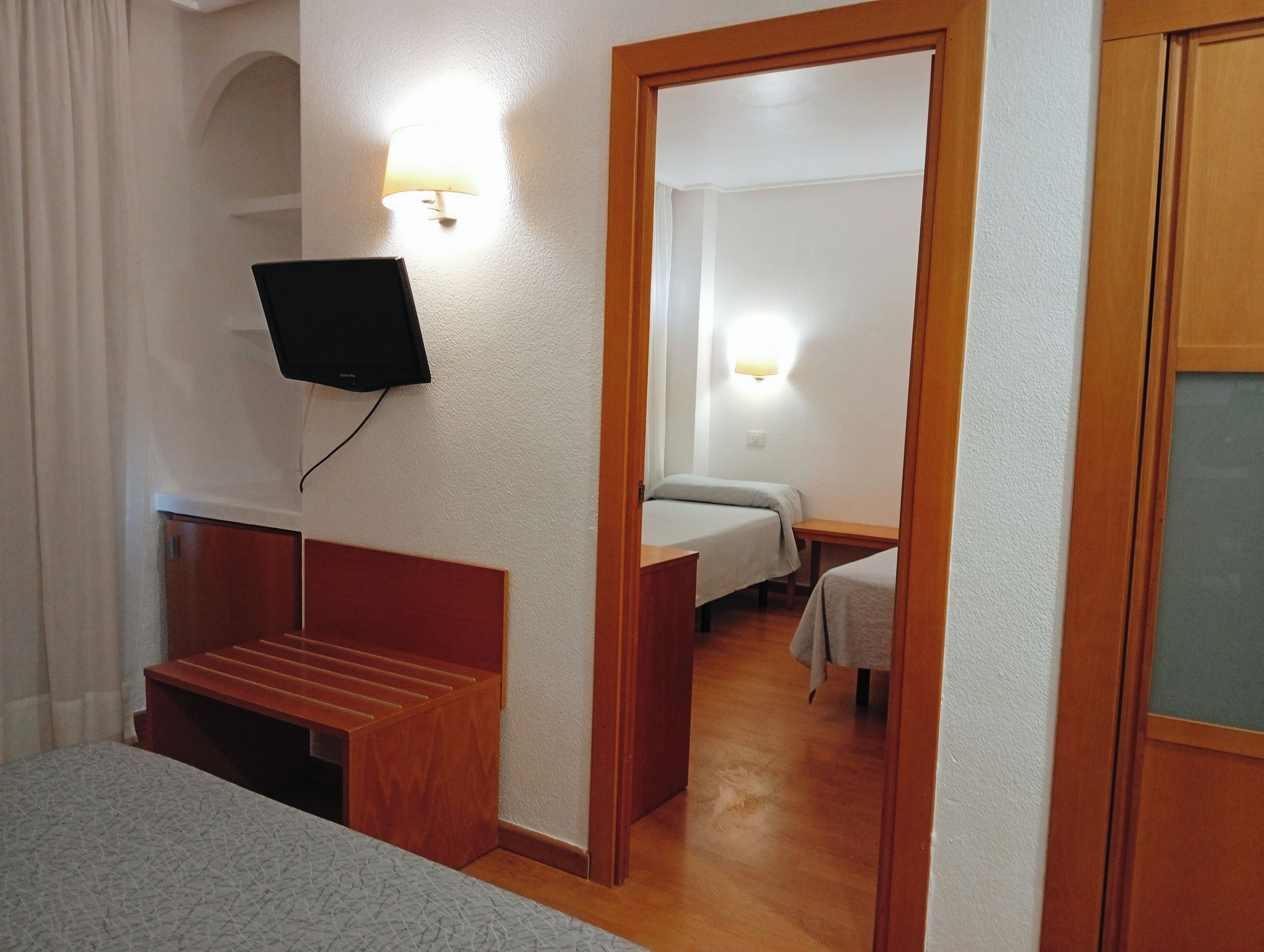 A hotel room with a wooden bench, a bed, a desk, and a TV mounted on the wall.
