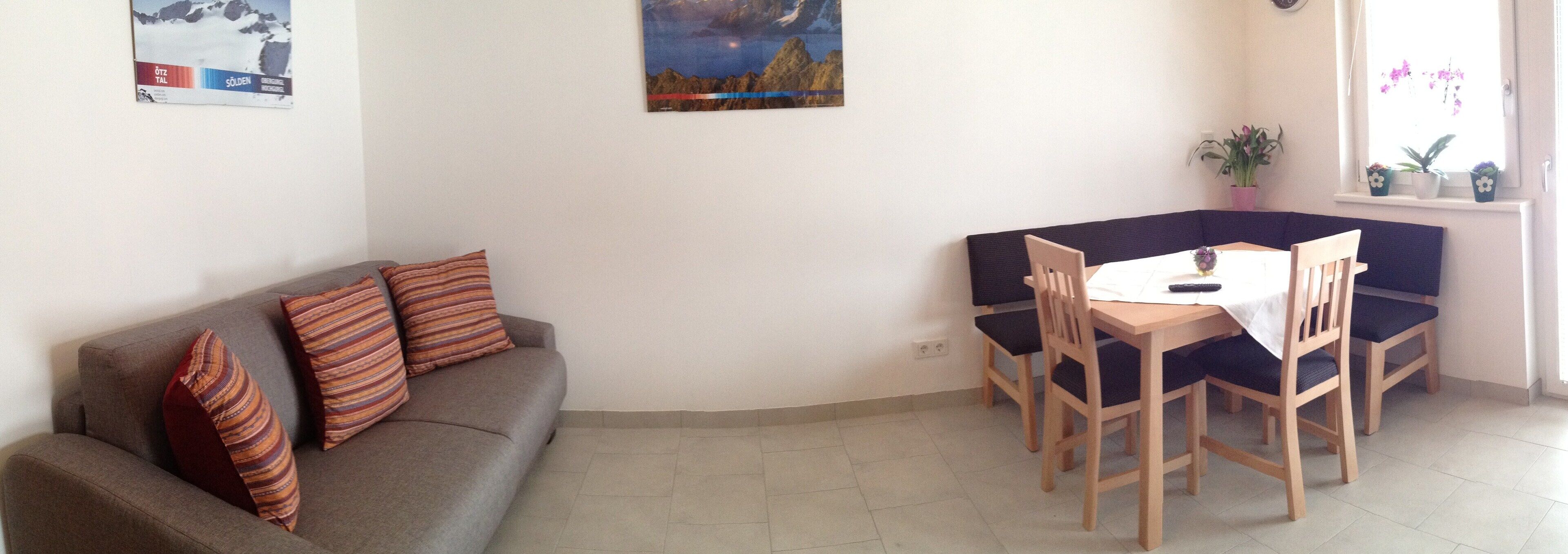 Een woonkamer met een bank, een eettafel met stoelen en een raam met planten.