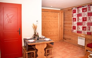 A dining area with a wooden table set for four, a red door with a floral design, and a wooden wall with a red and white patterned curtain.