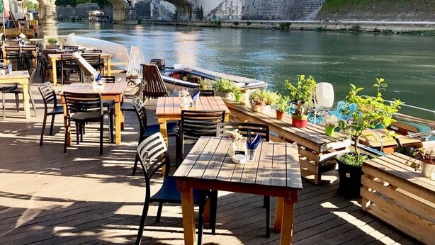 Zona pranzo all'aperto con tavoli e sedie, affacciata su un fiume. Si vedono un ponte e degli edifici sullo sfondo.