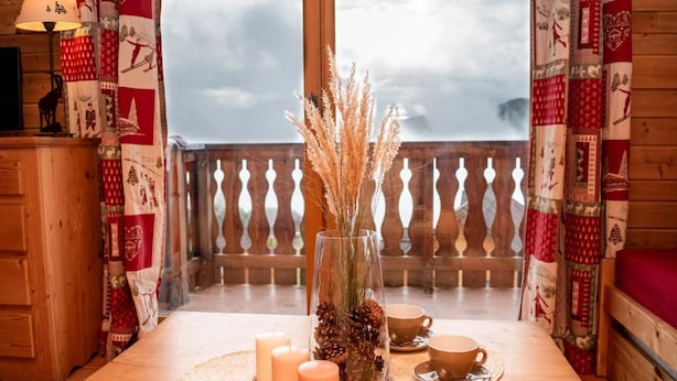 Une table en bois avec des bougies, un vase en verre contenant des pommes de pin et une tasse sur un balcon orné de rideaux à motifs.