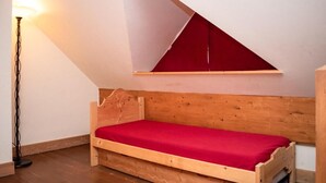 Un petit lit en bois avec un matelas rouge, dans une pièce dont le plafond est incliné et qui est éclairée par un lampadaire.