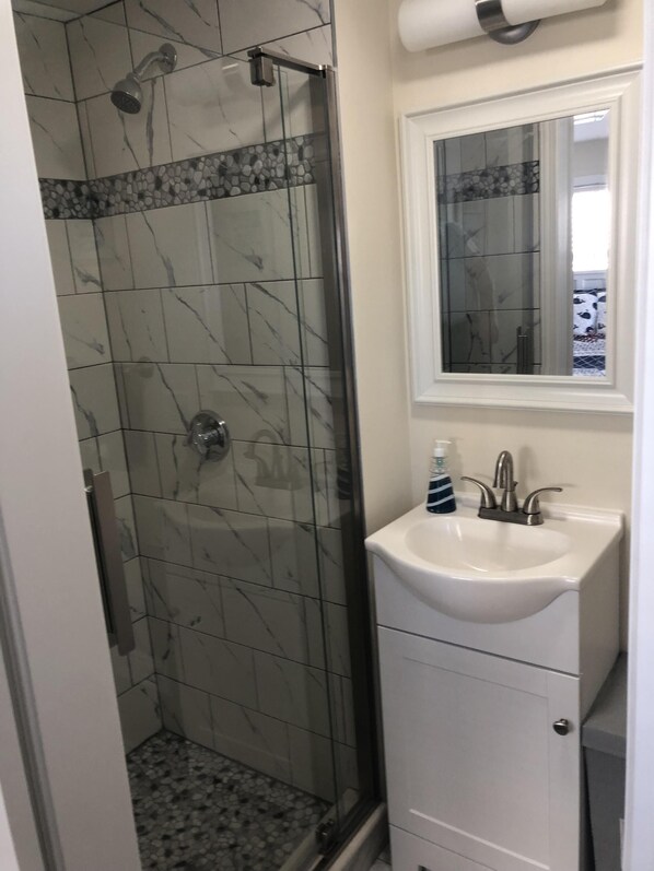 Un baño con ducha de vidrio, un lavabo blanco con espejo encima y suelo de baldosas.