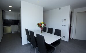 A modern dining area with a white table and black chairs, a vase with flowers, and a kitchen in the background. - New apartment with terrace and pool near the beach and shops Inmosantos Nuria Atico (Rosas / Roses)