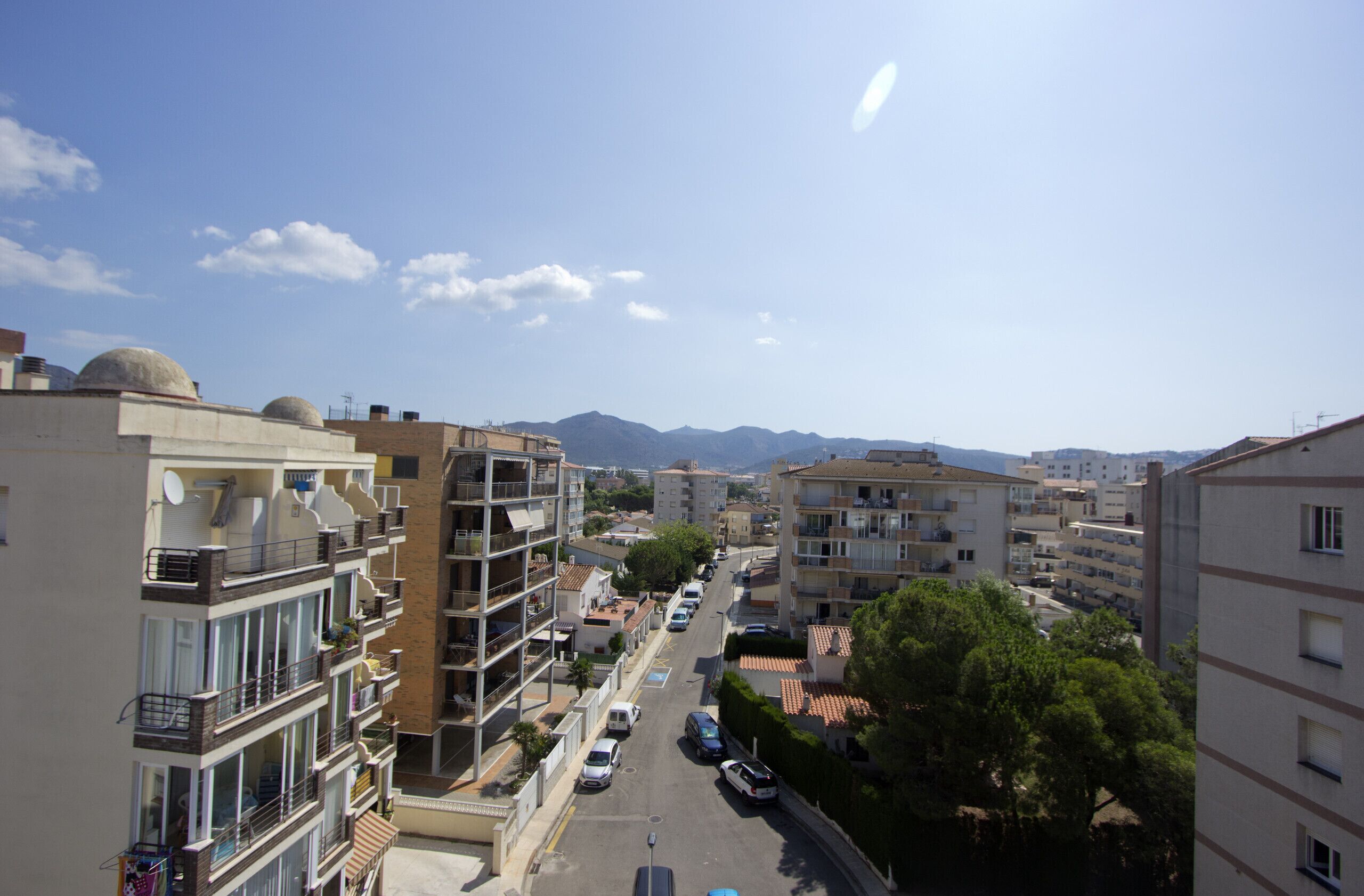 A multi-story residential building with balconies, a street in the foreground, and mountains in the background.