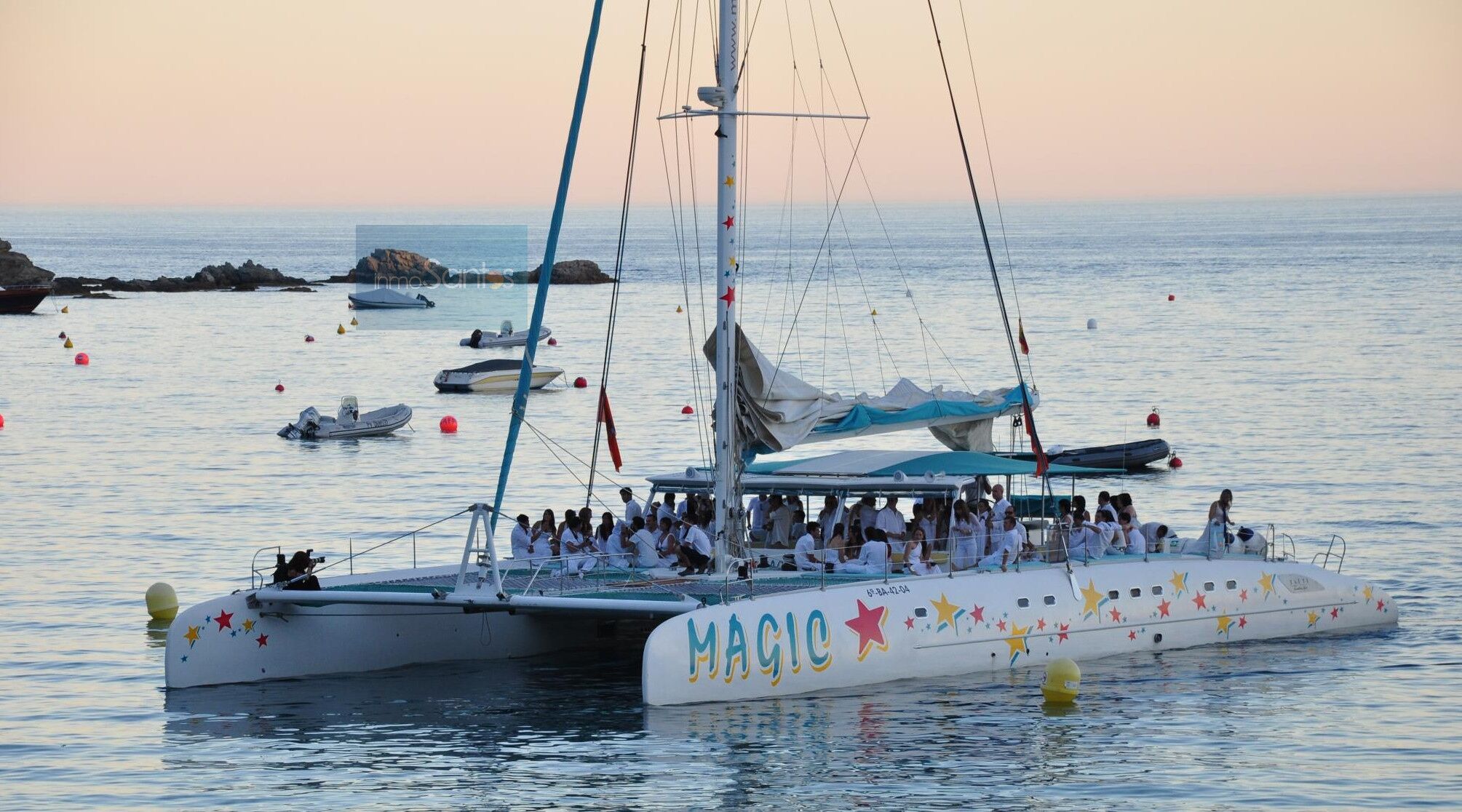 Um veleiro chamado MAGIC, com várias pessoas a bordo, atracado em uma marina, com outros barcos e boias ao fundo.