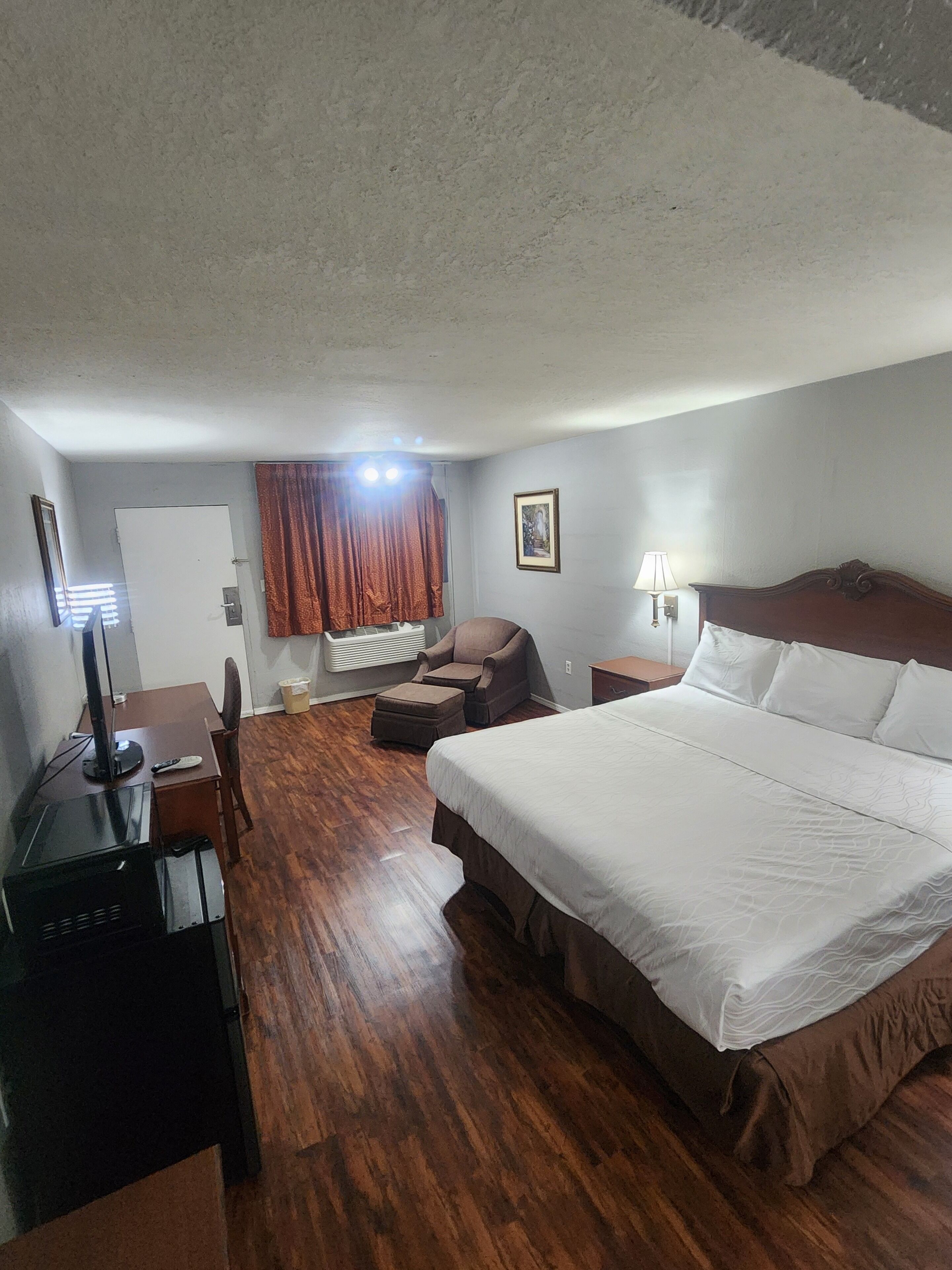 A hotel room with a large bed, a desk with a computer, a chair, a lamp, and a window with curtains.