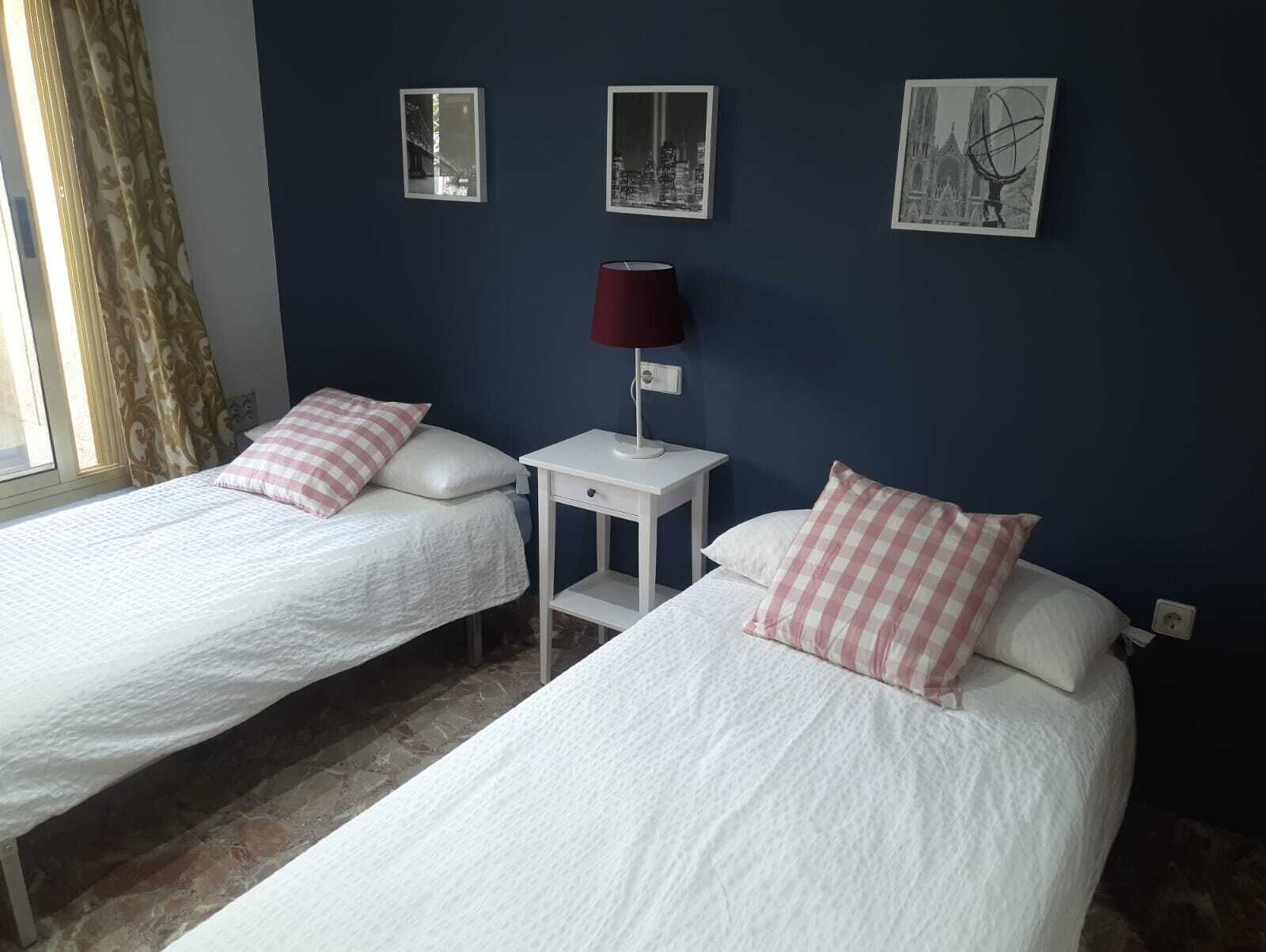 Una habitación con dos camas, cada una con ropa de cama blanca y almohadas a cuadros rosas. Hay una mesita de noche pequeña blanca con una lámpara roja entre las camas. Las paredes están pintadas de azul oscuro, y hay tres cuadros enmarcados colgados en la pared.