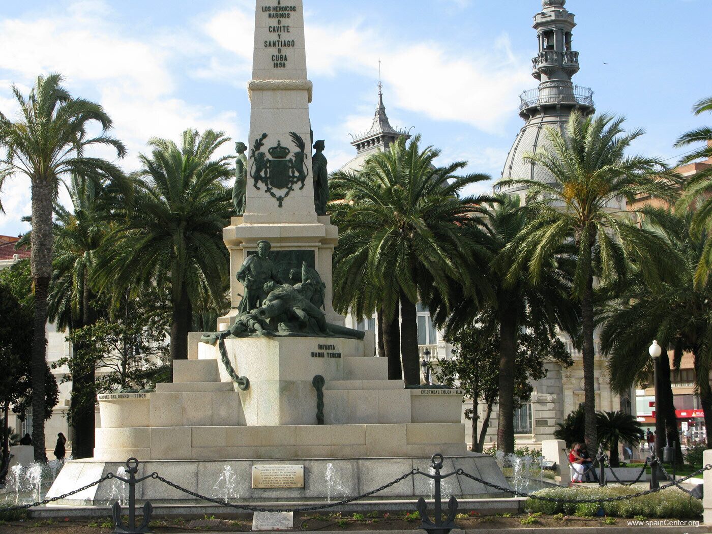 Um monumento com inscrições, estátuas e uma fonte numa praça pública, com palmeiras e um edifício ao fundo.