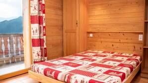 A wooden room with a bed, a window with a view of mountains, and a curtain with a pattern.