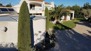 Ein modernes Haus mit Flachdach, Solaranlage, einem gepflegten Rasen und einer kleinen Gartenanlage mit einer Palme und Liegestühlen.